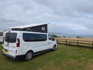 White Renault Trafic campervan with a raised pop-top roof at a grassy campsite overlooking the sea.